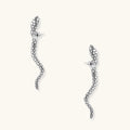 Silver-toned serpent-shaped dangle earrings with detailed scale texture curving in S-shape on beige background